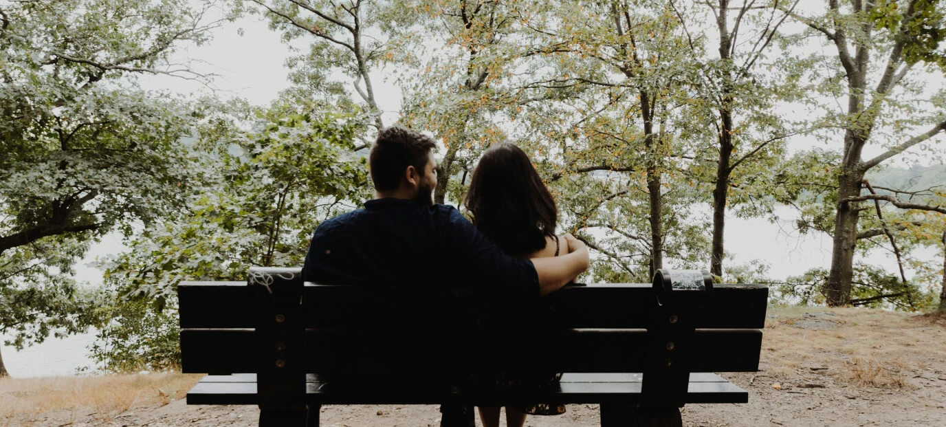 A couple seated on a park bench under a canopy of trees, overlooking a lake, symbolizing companionship and tranquility.
